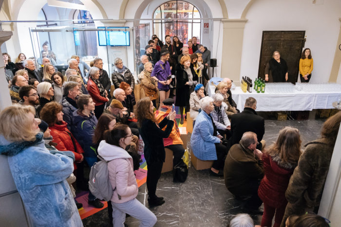 Eine Menschenmenge hört einem Redner in einem Saal zu - normal anders, mit Getränken auf einem Beistelltisch.