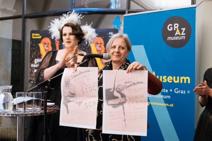 Zwei Frauen an Mikrofonen, eine mit zwei ganz normal,anderen Plakaten, bei einer Veranstaltung des Graz Museums.