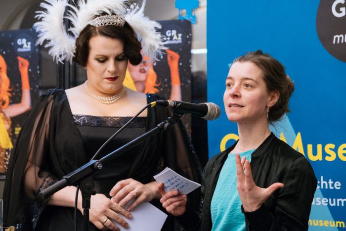 Zwei Personen an einem Mikrofon; ganz normal anders, einer liest von einer Karte ab, der andere gestikuliert.