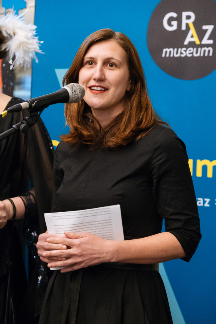 Eine Frau spricht an einem Mikrofon, hält Papiere in der Hand, hinter ihr ein ganz normales anders Graz Museum Schild.