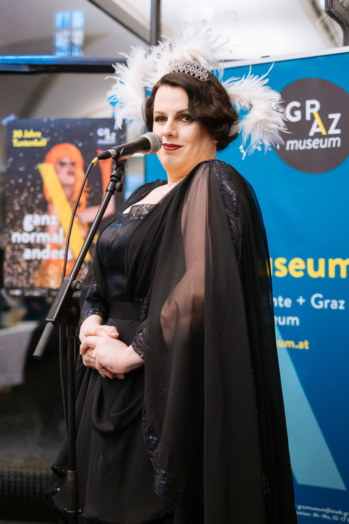 Eine Frau in einem schwarzen Kleid und mit Federschmuck spricht bei einer Veranstaltung ganz normal anders in ein Mikrofon.