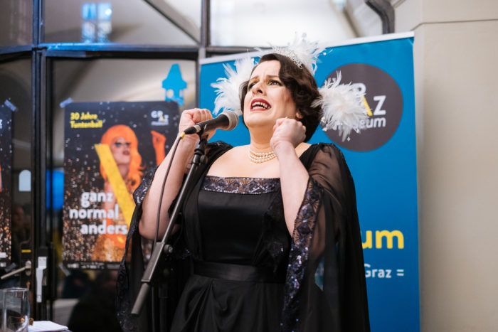 Frau in schwarzem Kleid und gefiedertem Kopfschmuck singt ganz normal und doch anders in ein Mikrofon.