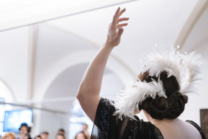 Person mit gefiederter Kopfbedeckung hebt den Arm-ganz normal anders- in einem Innenbereich.