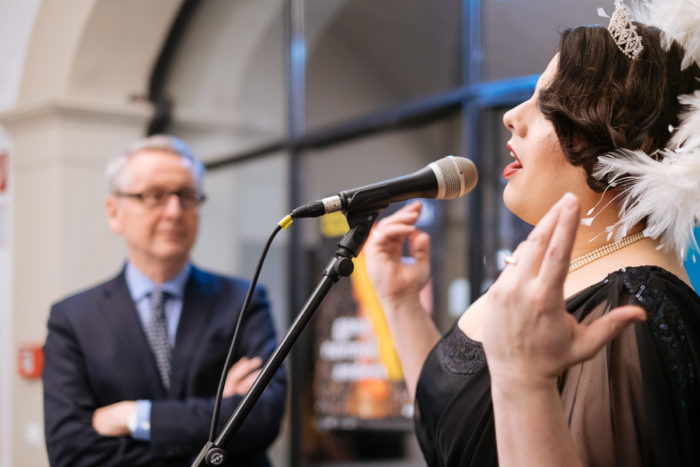 Eine Frau spricht oder singt in ein Mikrofon - das ist ganz normal, während ein Mann im Hintergrund steht.