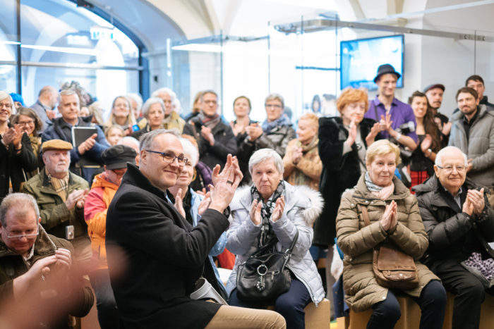 Eine Gruppe, ganz normal anders, sitzt und steht drinnen, klatscht und beobachtet die Front.