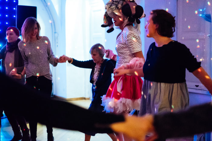 Eine Gruppe von Menschen, ganz normal anders, hält sich an den Händen und tanzt unter bunten Partylichtern in einem Haus.