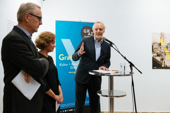 Drei Personen stehen bei einer offiziellen Veranstaltung in der Nähe eines Podiums, hinter ihnen ein blaues Boulevards-Banner.