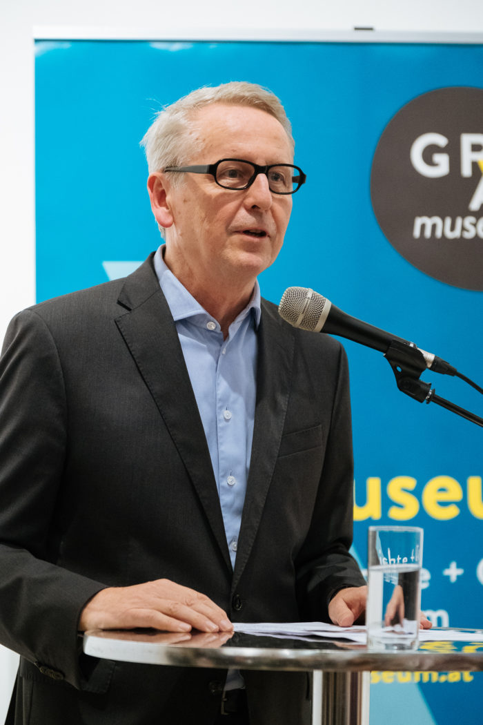 Mann im Anzug spricht an einem Podium mit Mikrofon; auf blauem Hintergrund ist der Text Brücken und Boulevards zu sehen.