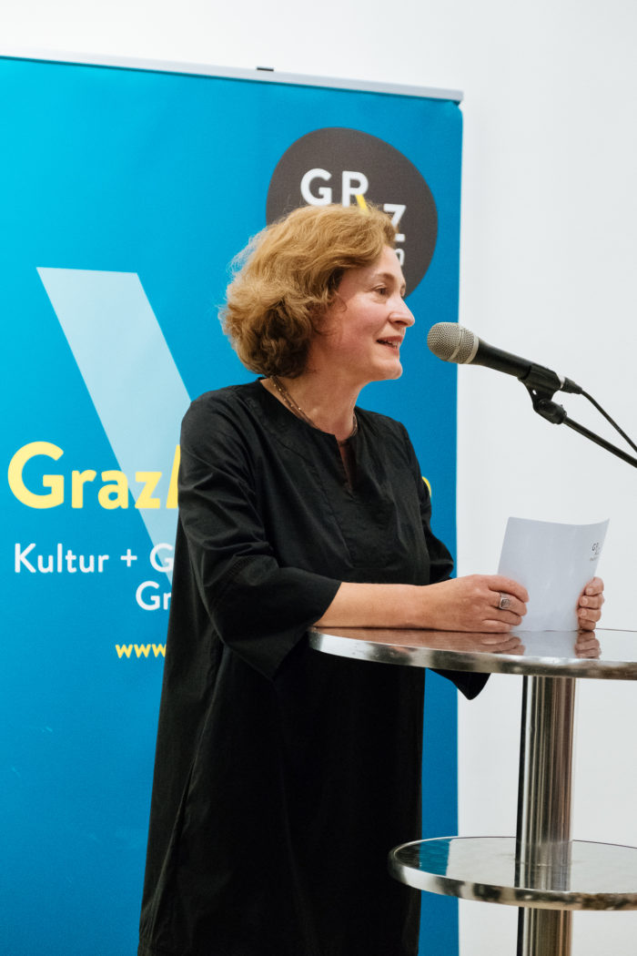 Eine Frau spricht an einem Podium mit Papieren; hinter ihr hängt ein blaues Brücken-Banner.