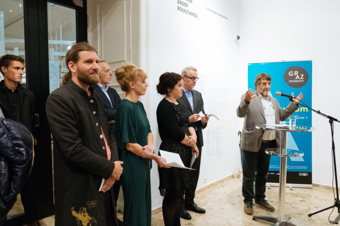 Eine Gruppe von Menschen steht in einem Innenraum, während ein Mann auf einem Podium während einer Veranstaltung spricht.