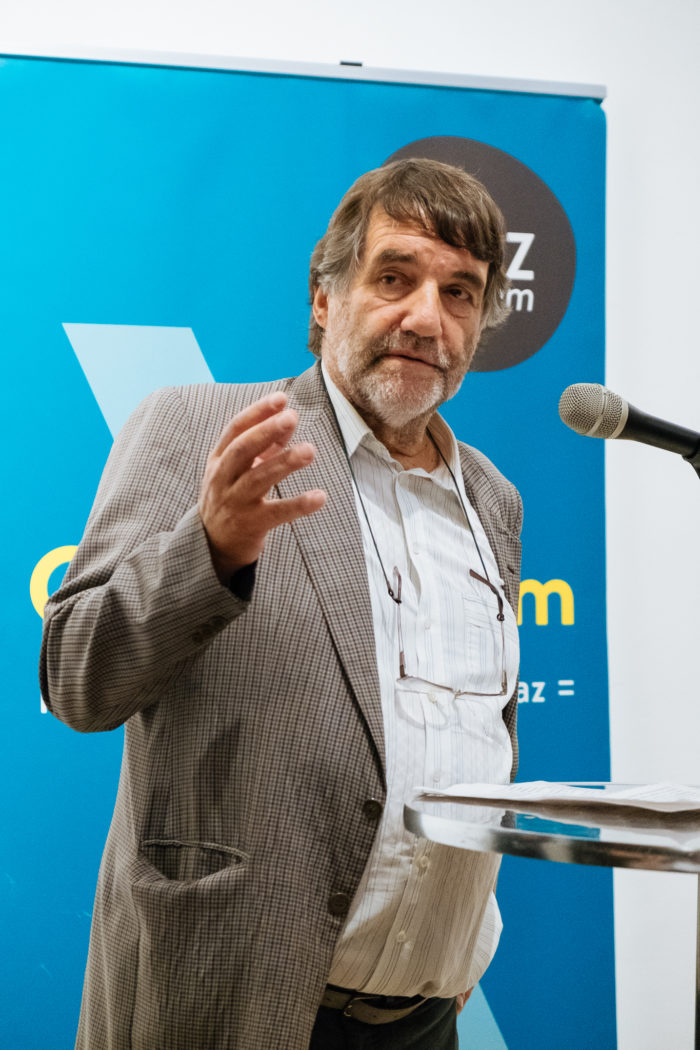 Ein älterer Mann spricht an einem Podium mit einem Mikrofon und einem blauen Banner im Hintergrund.