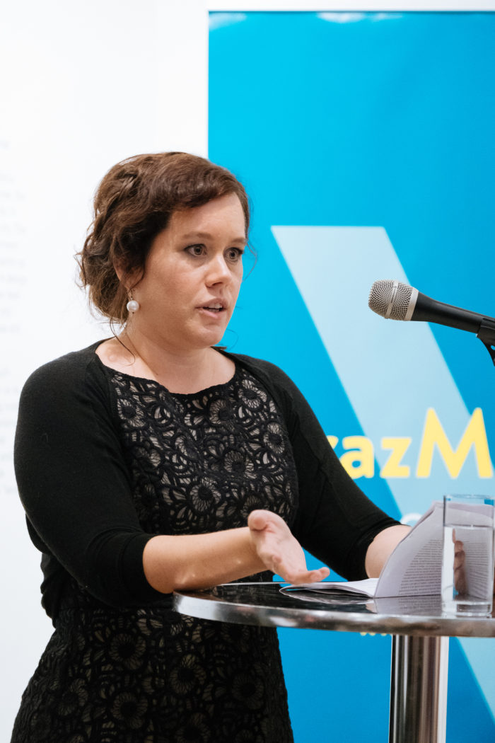 Frau spricht an einem Podium mit Mikrofon und Papieren, blaues Schild im Hintergrund.