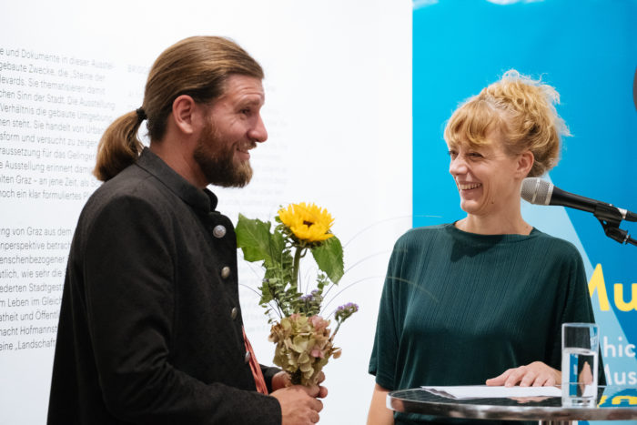 Ein Mann mit Blumen steht neben einer lächelnden Frau, die in ein Mikrofon spricht.