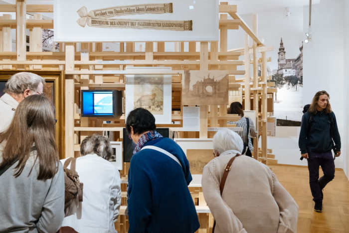 Besucher betrachten Exponate und gerahmte Kunstwerke in einem Museum mit hölzernen Boulevard-Strukturen.