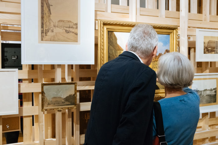 Ein älteres Ehepaar betrachtet gerahmte Kunstwerke in einer Galerie mit Holzgitterwänden und Boulevardszenen.