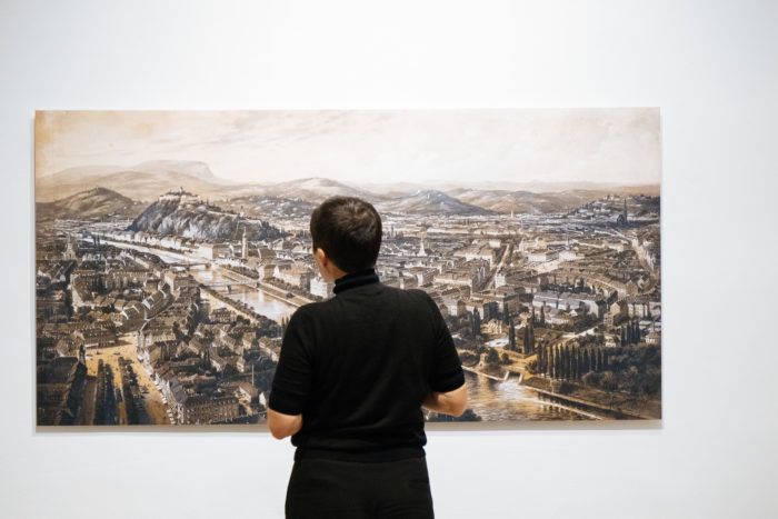 Eine Person steht vor einer großen Boulevardlandschaft, die auf einer weißen Galeriewand dargestellt ist.