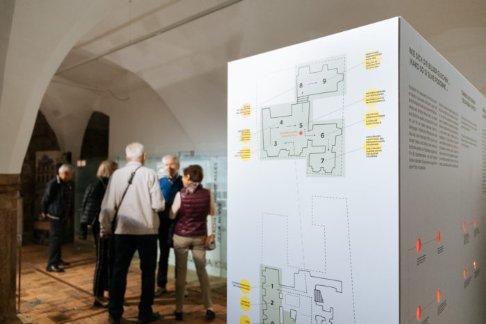 Eine Gruppe von Personen steht in einem Ausstellungsraum neben einem Informationsdisplay mit Grundrissen.