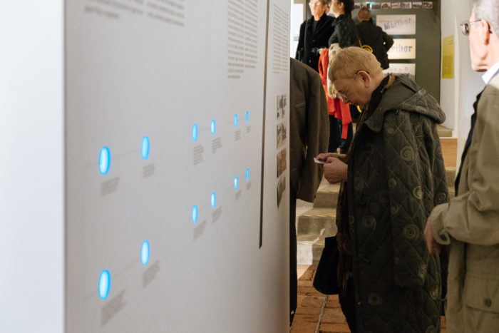 Betrachter eines Exponats mit blauen Leuchtpunkten und Text auf einer weißen Ausstellungswand.