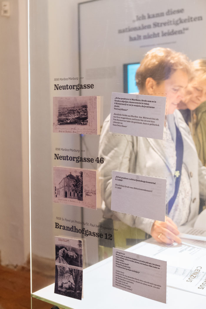 Zwei Personen lesen Text neben beschrifteten historischen Fotos und Beschreibungen auf einer Glasplatte.