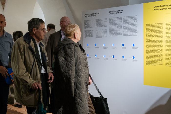 Mehrere Personen lesen aufmerksam die Informationstafeln, die an einer Museums- oder Ausstellungswand angebracht sind.