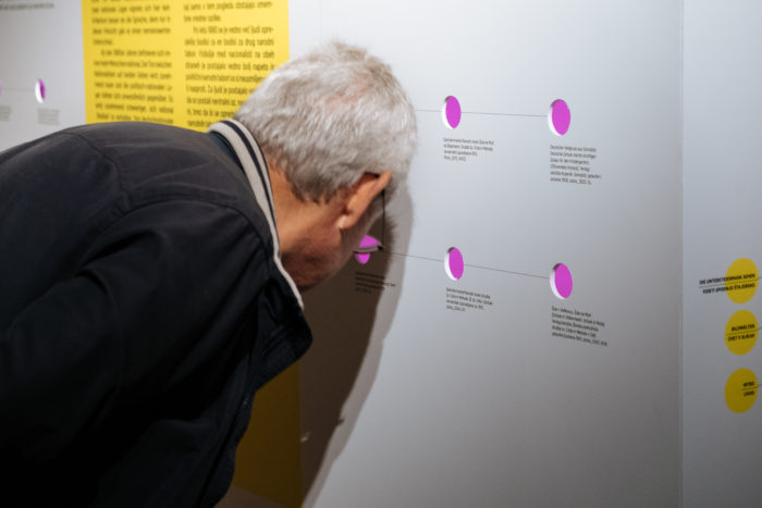 Ein älterer Mann betrachtet aufmerksam ein Display mit Text und Diagrammen an einer weißen Wand.