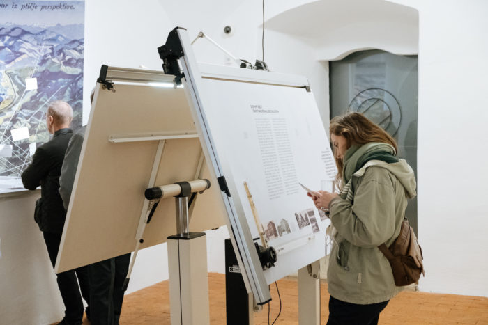 Frau schaut auf ihr Telefon neben einer großen Anzeigetafel in einem Ausstellungsraum.