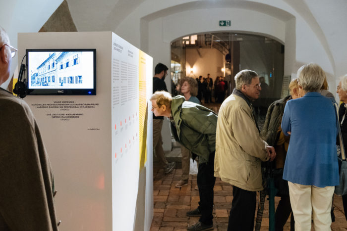 Menschen betrachten ein Ausstellungsstück in einer Galerie; eine Person schaut sich den Text auf einer Schautafel genau an.