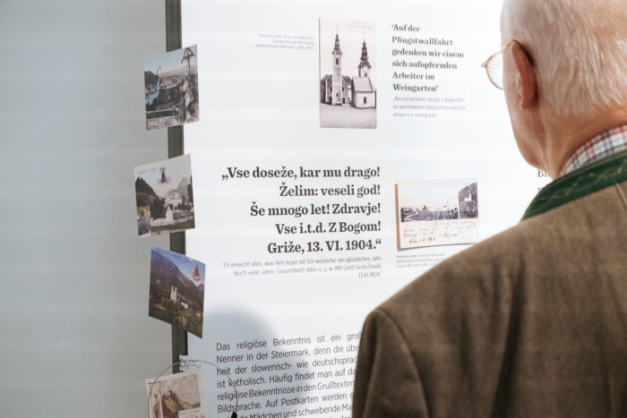 Ein älterer Mann schaut auf eine Schautafel mit alten Fotos und Texten in slowenischer und deutscher Sprache.