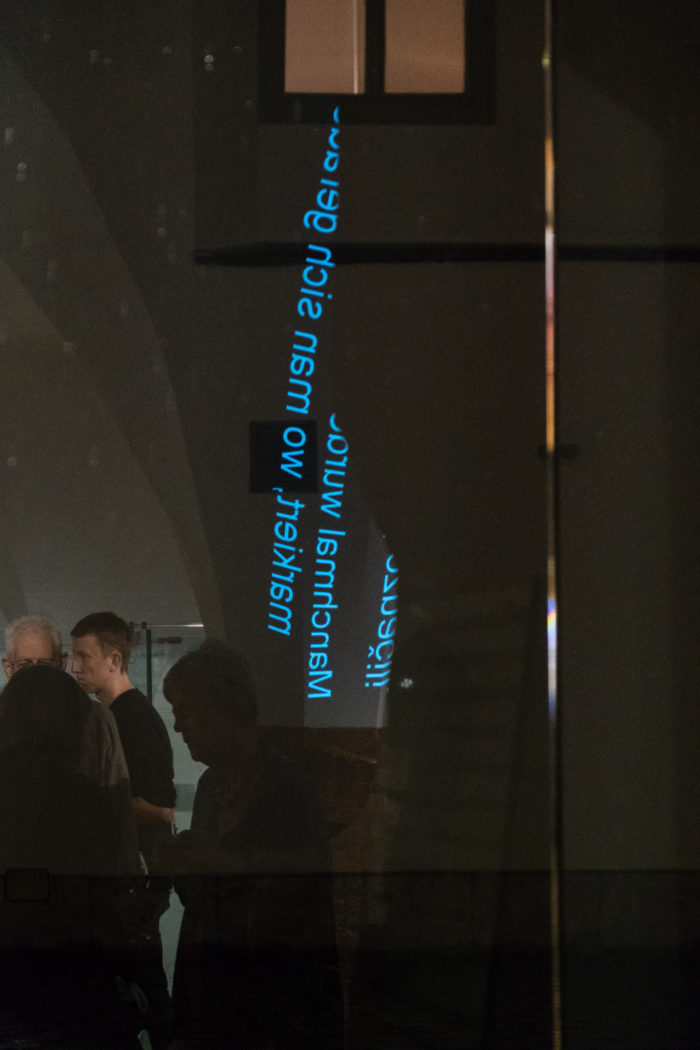Schwach beleuchteter Raum mit Menschen als Silhouetten; blauer Text spiegelt sich auf einer Glasfläche.