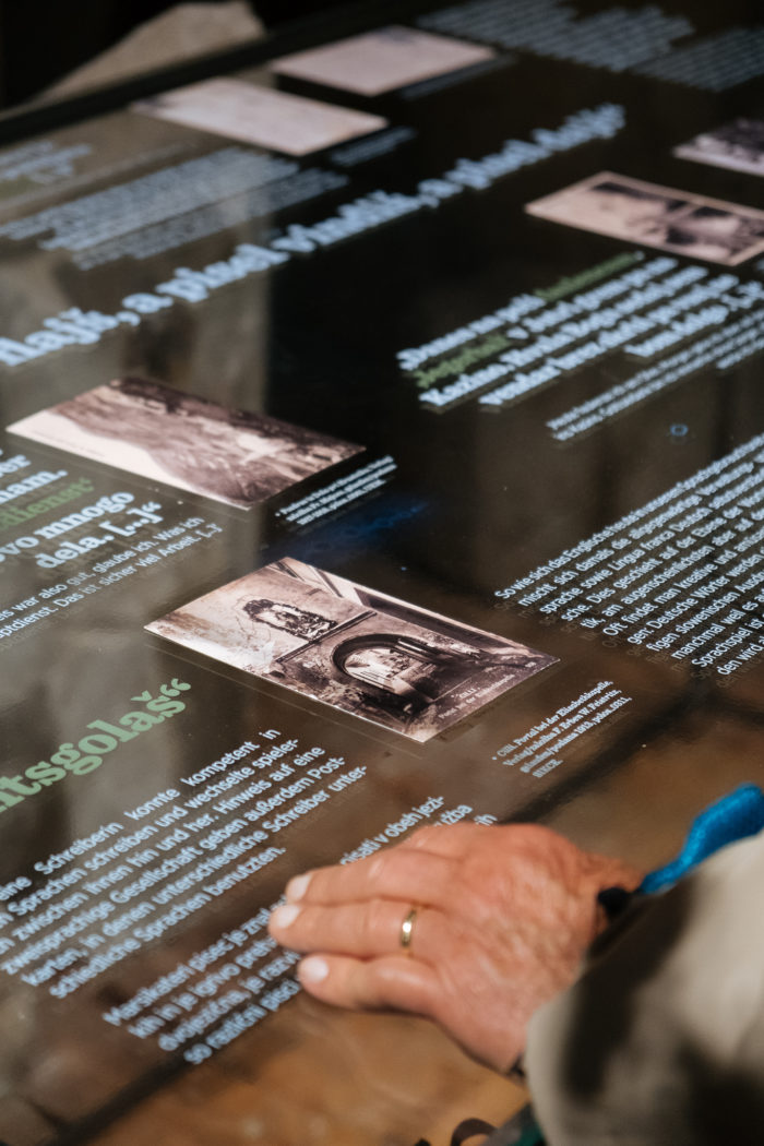 Eine Person liest Text und betrachtet historische Fotos auf einer Glastafel.