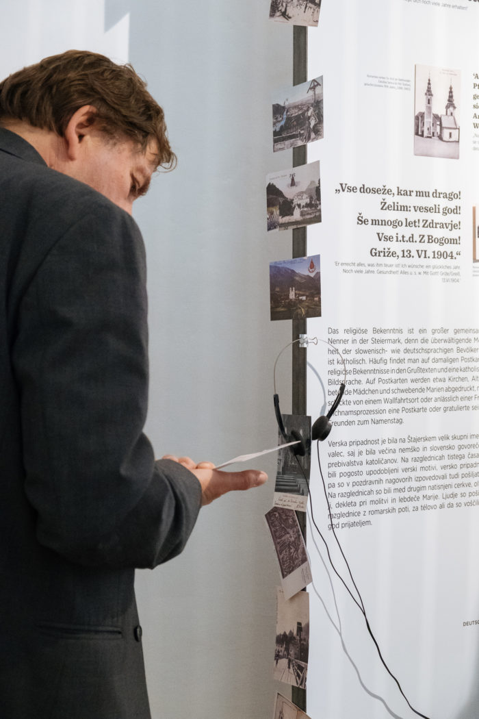 Ein Mann betrachtet Fotos und Texte, die auf einer vertikalen Ausstellungstafel mit Kopfhörern angezeigt werden.