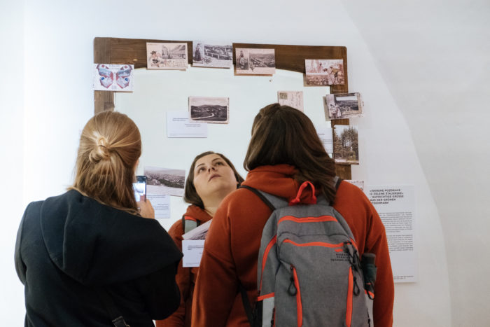 Drei Personen betrachten Fotos und Notizen an einer Wand, eine macht ein Foto mit dem Handy.