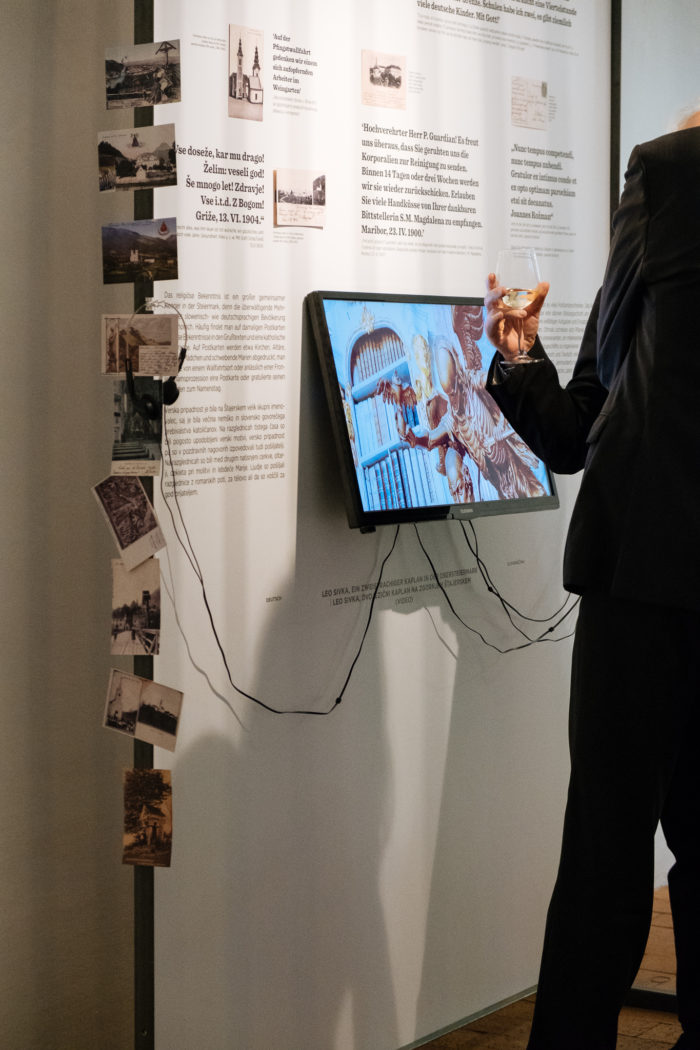 Eine Person mit einem Getränk steht in der Nähe einer multimedialen Museumsausstellung mit Bildern und Texten an einer Wand.