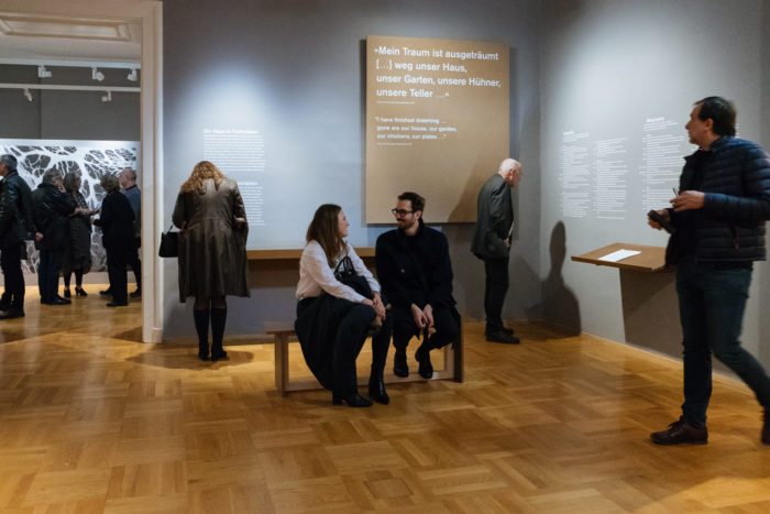 Menschen betrachten Exponate auf dem großen Bruch; zwei ruhen sich auf einer Museumsbank in der Mitte der Galerie aus.