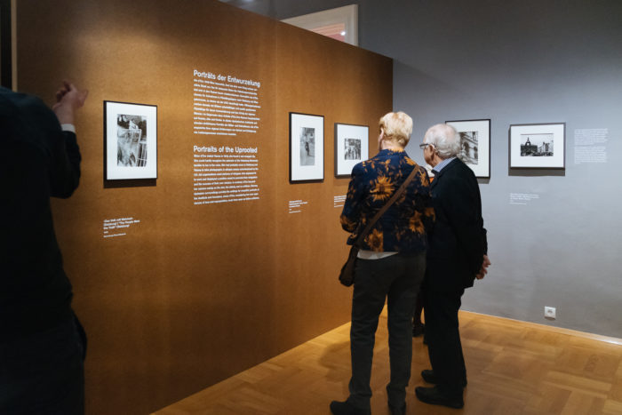 Zwei Personen betrachten den Text und die gerahmten Fotos der Ausstellung Großer Bruch an einer braunen Galeriewand.