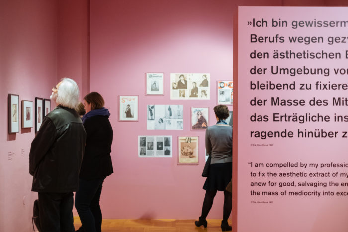 Drei Personen betrachten gerahmte Kunstwerke an einer rosafarbenen Wand und erkunden das Thema des großen Bruchs in einer Galerie.