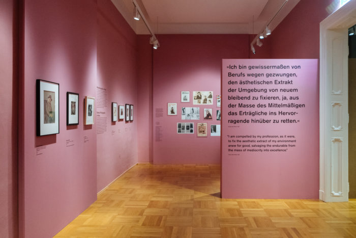 Kunstgalerie mit rosafarbenen Wänden, gerahmten Kunstwerken und einer großen Texttafel zum Großen Bruch in deutscher und englischer Sprache.