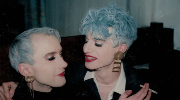 Zwei Personen mit blauen Haaren und auffälligem Make-up sitzen eng beieinander und erinnern an den Stil der 1990er-Jahre in Graz.
