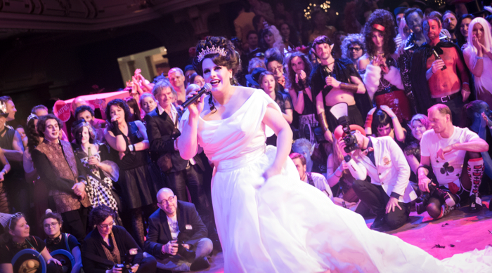 Miss Alexandra Desmond in einem weißen Kleid und einer Krone singt auf der Bühne und begeistert das kostümierte Publikum.