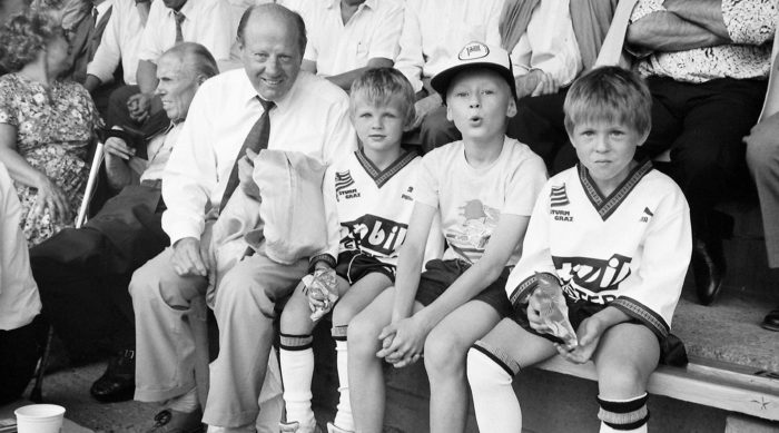 Ein älterer Mann sitzt mit drei Jungen in Sportuniformen in der Gruabn-Kinderwerkstatt.