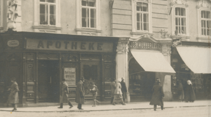 Menschen, die in dieser Straßenchronik des Jahrgangs 1936 an Geschäften, darunter eine Apotheke, vorbeigehen.