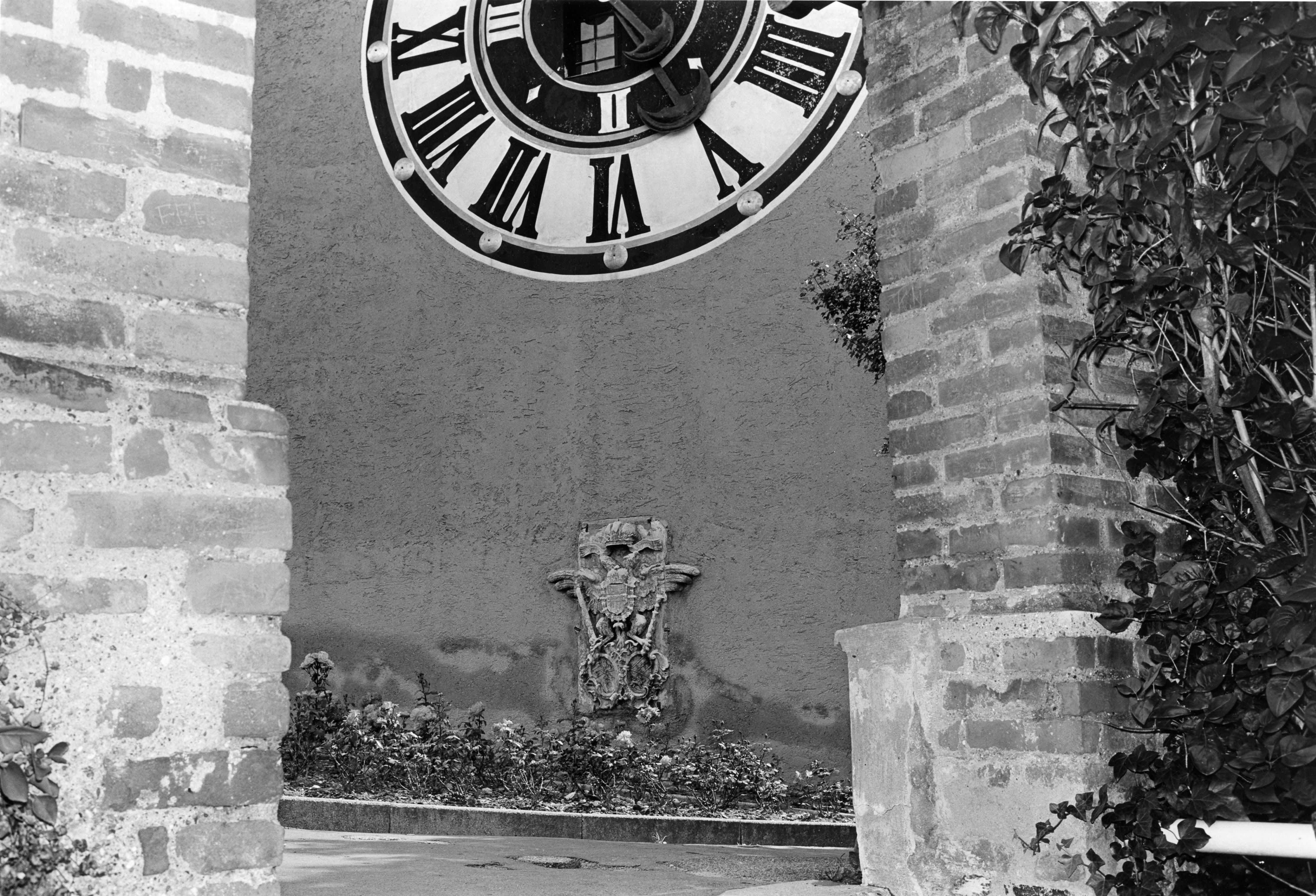 Große SCHLOSSBERG-Uhr mit römischen Ziffern über einer Steinskulptur, eingerahmt von Backsteinsäulen.