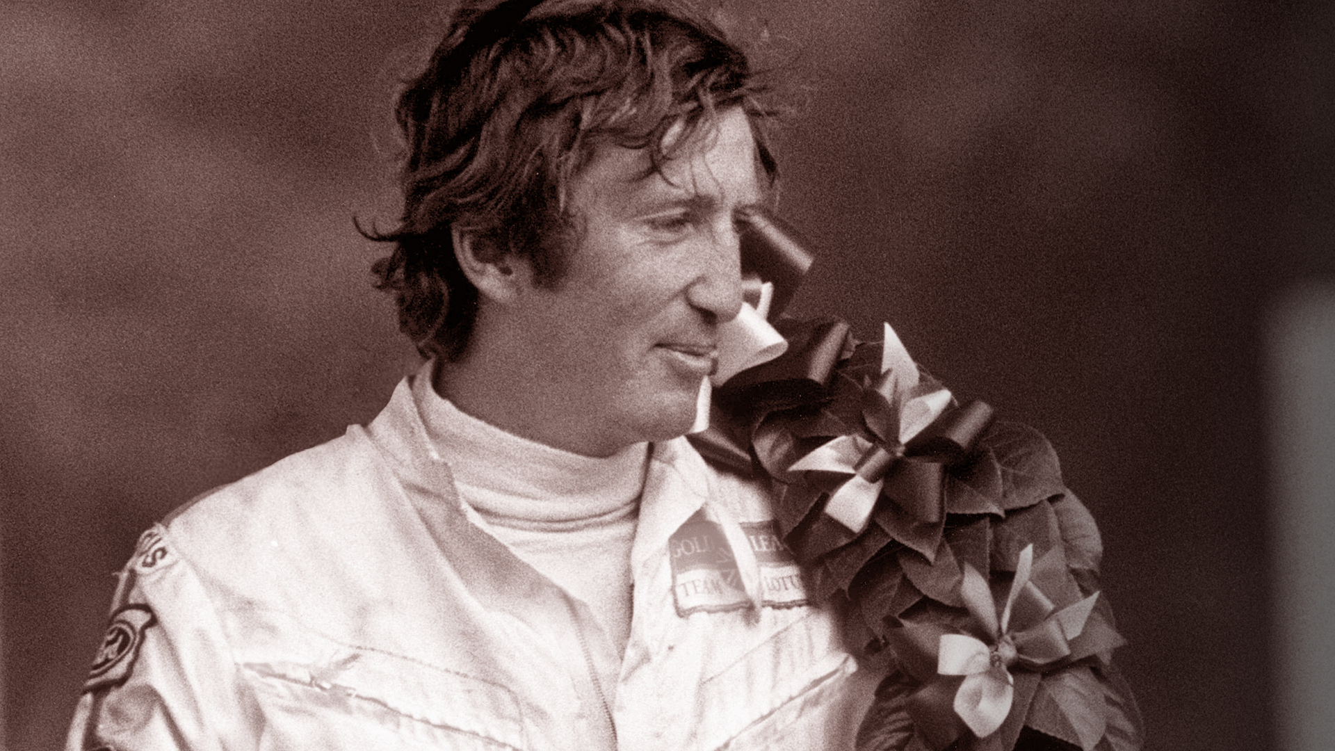 Ein weiß gekleideter Rennfahrer, der den Mythos von Jochen Rindt verkörpert, steht da und lächelt.