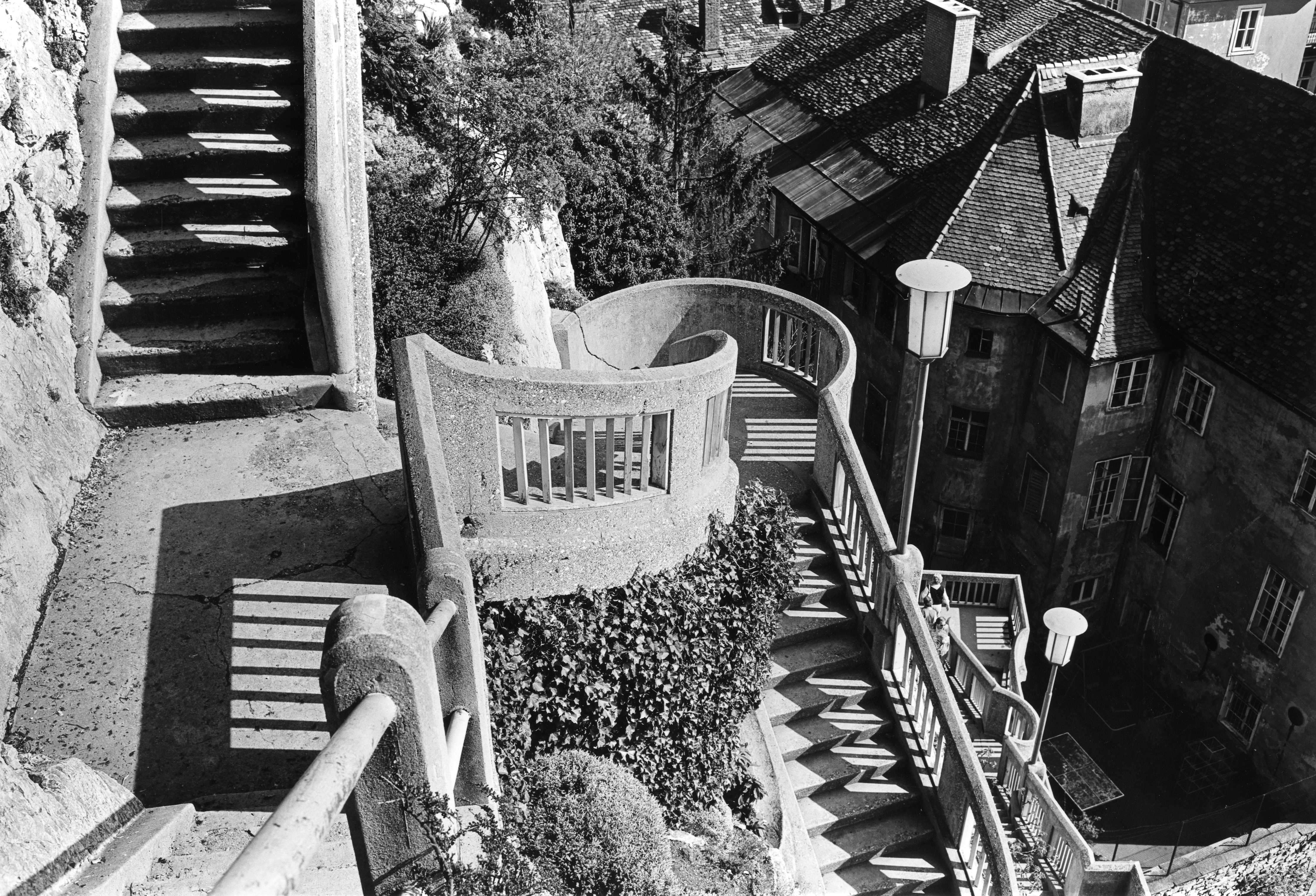 Eine geschwungene Betontreppe führt neben alten SCHLOSSBERG-Gebäuden und Laub im Sonnenlicht hinunter.