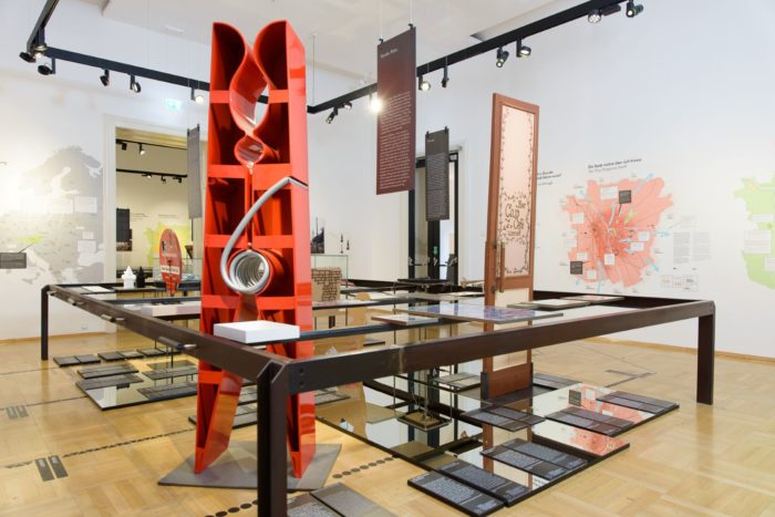In einem Raum in der Ausstellung steht im Vordergrund eine große, rote Wäscheklammer. Im Hintergrund ist eine historische IEngangstür zu sehen. Am Boden sind Spiegelflächen und Tafeln mit Texten montiert. An den Wänden gibt es Landkarten.