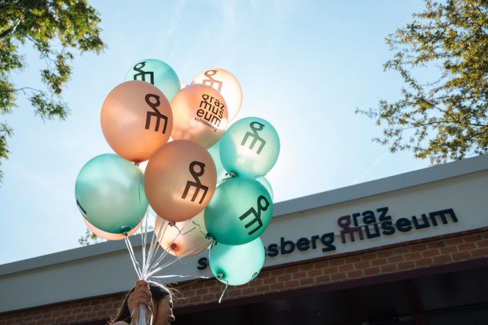 Person mit blaugrünen und orangefarbenen Luftballons in der Nähe des Graz Museums mit dem Schlossberg im Hintergrund.