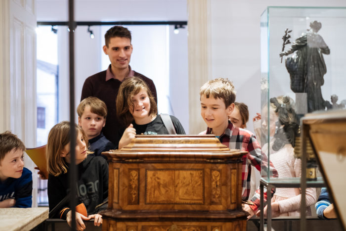 Kinder und ein Erwachsener erkunden eine Holzvitrine in der Ausstellung des Junge Stadt Ferienworkshops.