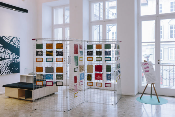 Farbenfrohe Drucke von Kindern erhellen Drahttafeln in einer sonnigen Grazer Galerie mit großen Fenstern.