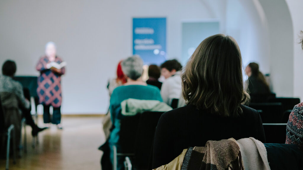 In einem Raum sitzende Menschen hören zu, während sich vorne die Lange Nacht der Genderlesung entfaltet.