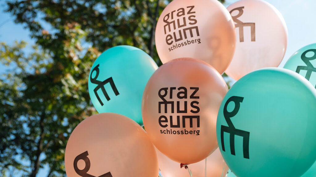 Pfirsichfarbene und blaugrüne Luftballons mit dem Logo des Museums Schlossberg in Graz.
